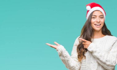 Hayret ve el ile sunulması ve parmak ile işaret ederken kameraya gülümseyen izole arka plan üzerinde Noel şapka giyen genç Arap kadın.