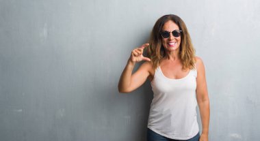 Orta yaş İspanyol kadın gri duvar gülümseyen güneş gözlüğü ve süre seyir ve kamera boyutu işaret parmaklarıyla yapıyor el ile kendine güvenen el hareketi yüzünden. Ölçü kavramı.