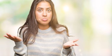 İzole arka plan clueless ve şaşkın bir ifadeyle kollar ve eller kaldırdı üzerinde kış kazak giyen genç güzel Arap kadın. Şüphe kavramı.