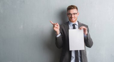 Genç redhead iş adamı boş kağıt tutan gri grunge duvarın üzerinden sözleşme çok mutlu el ve parmak ile işaret yan