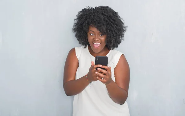 Young african american woman over grey grunge wall texting a message using smartphone scared in shock with a surprise face, afraid and excited with fear expression