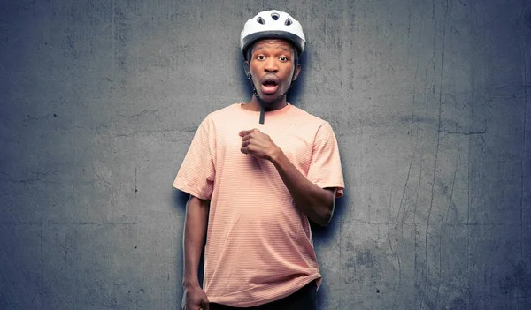 Black man wearing bike helmet happy and surprised cheering expressing wow gesture, pointing with finger