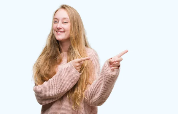 ブロンド十代の若者の女性は身に着けているピンクのセーター笑顔と側に つの手と指で指しているカメラ目線 — ストック写真