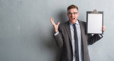 Genç redhead iş adamı ile çığlık zaferini kutluyor Pano çok mutlu ve heyecanlı, kazanan ifade büyük holding gri grunge duvarın üzerinden gülümse ve el kaldırdı