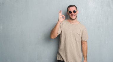 Beyaz delikanlı el ve parmaklar ok işaretiyle yapıyor olumlu gülümseyen güneş gözlüğü takıyor gri grunge duvarın üzerinden. Başarılı ifade.