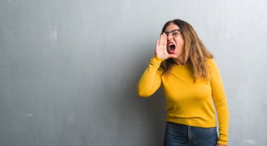 Orta yaş İspanyol kadın bağırarak ve ağzı üzerinde el ile yan için çığlık gözlüklü gri duvar yüzünden. İletişim kavramı.