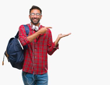 Hayret ve el ile sunulması ve parmak ile işaret ederken kameraya gülümseyen izole arka plan üzerinde kulaklık ve sırt çantası giyiyor yetişkin İspanyol öğrenci adam.