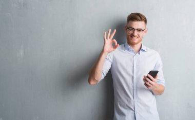 Genç Kızıl saçlı adam gri grunge duvar parmak, mükemmel sembolü ile ok işareti yapıyor smartphone kullanarak ileti gönderme