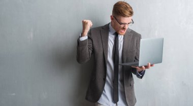 Genç redhead zarif iş adamı dizüstü kullanarak gri grunge duvarın üzerinden rahatsız ve boşa çıkarmak ile öfke, çılgın da yükseltilmiş el ile bağırmaya kavramı öfke