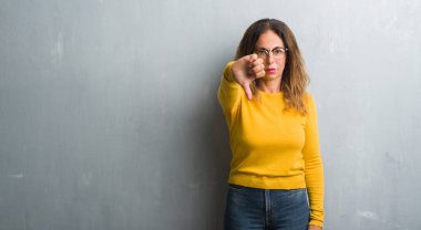 Orta yaş İspanyol kadın mutsuz ve kızgın ret gösterilen ve başparmak aşağı hareketi ile negatif seyir gözlüklü gri duvar yüzünden. Kötü ifade.