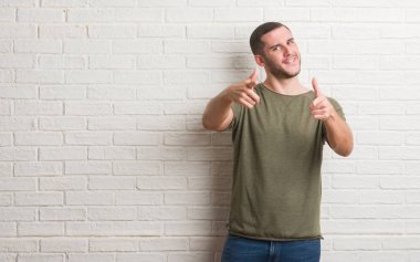 Fotoğraf makinesi ile mutlu ve komik yüz için parmakları işaret beyaz tuğla duvarın üzerinden ayakta beyaz delikanlı. İyi enerji ve vibes.