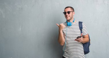 Gri grunge duvar işaret ve başparmak kadar mutlu yüz gülümseyen tarafı ile gösterilen smartphone kullanarak kulaklık giyen genç beyaz öğrenci adam