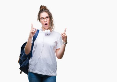 Sırt çantası ve kulaklık izole arka plan üzerinde giyen genç esmer öğrenci kız hayran ve yukarıya bakıp parmakları ile işaret şaşırttı ve silah kaldırdı.