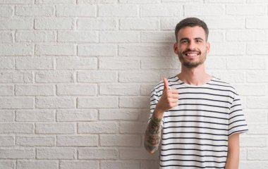Genç yetişkin adam beyaz tuğla duvardan mutlu büyük gülümseme ok işareti, başparmak ile parmak, mükemmel işareti yapıyor ayakta