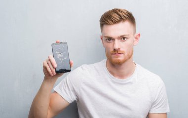 Genç Kızıl saçlı adam kendinden emin bir ifade ile kırık smartphone ciddi düşünme acımak yüz tutan gri grunge duvarın üzerinden
