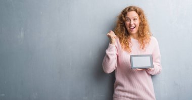 Gri grunge duvar kullanarak üzerinde genç Kızıl saçlı kadın tablet gurur çığlık ve zafer ve başarı çok heyecanlı, kutluyor duygu tezahürat