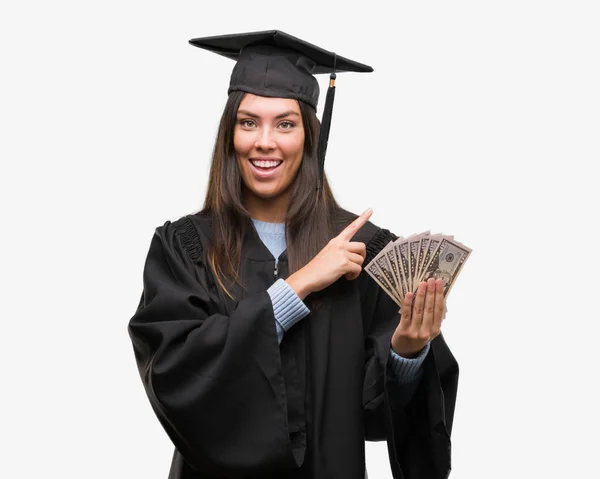 着た若いヒスパニック系女性卒業制服持株ドル側の手と指で指す非常に満足 — ストック写真