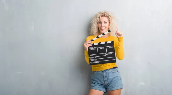 Young Blonde Woman Grunge Grey Wall Holding Calpboard Movie Surprised — Stock Photo, Image