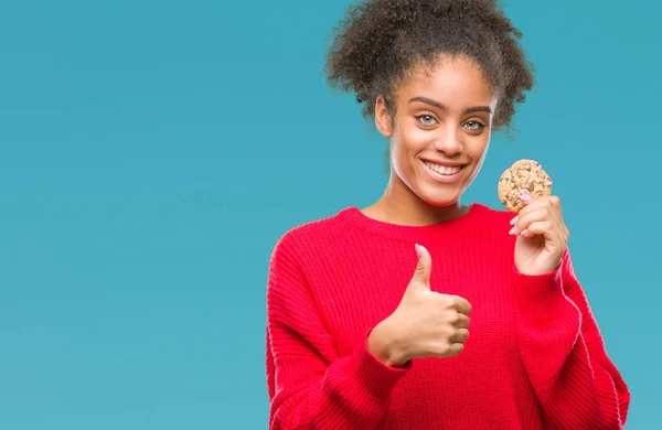 Young afro american woman eating chocolate cookie over isolated background happy with big smile doing ok sign, thumb up with fingers, excellent sign