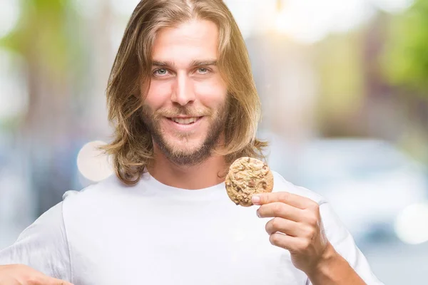 驚きと孤立した背景にチョコレート クッキーを食べて長い髪のハンサムな若者に直面する自分自身に人差し指 — ストック写真