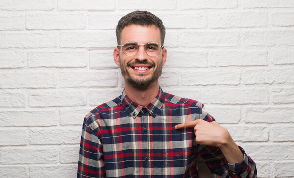 Young adult man standing over white brick wall with surprise face pointing finger to himself