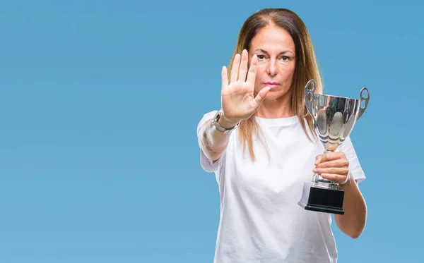 Middle age hispanic winner woman celebrating award holding trophy over isolated background with open hand doing stop sign with serious and confident expression, defense gesture