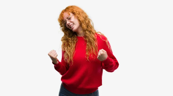 赤毛の若い女性非常に幸せと興奮の腕を上げ 勝者のジェスチャを行う赤いセーターを着て笑顔で成功のために叫んで お祝いのコンセプト — ストック写真