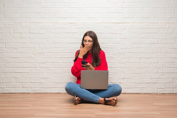 年轻的黑发女子坐在地板上使用膝上型电脑和智能手机捂住嘴用手震惊的错误 表达恐惧 害怕沉默 秘密概念 — 图库照片