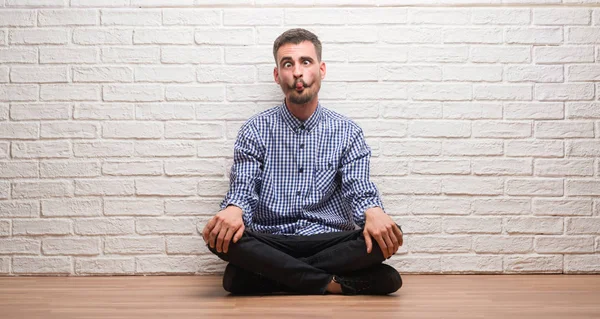 Young adult man sitting over white brick wall making fish face with lips, crazy and comical gesture. Funny expression.