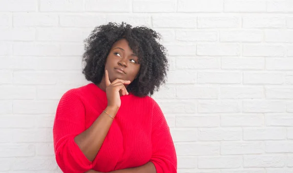 Young african american plus size woman over white brick wall with hand on chin thinking about question, pensive expression. Smiling with thoughtful face. Doubt concept.