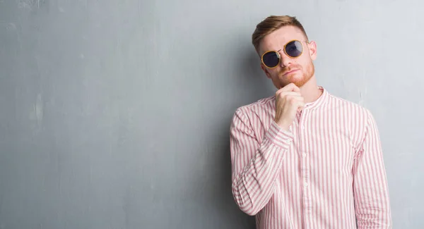 Young redhead man over grey grunge wall wearing retro sunglasses serious face thinking about question, very confused idea