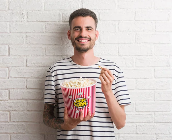 stock image Young adult man eating popcorn standing over white brick wall with a happy face standing and smiling with a confident smile showing teeth