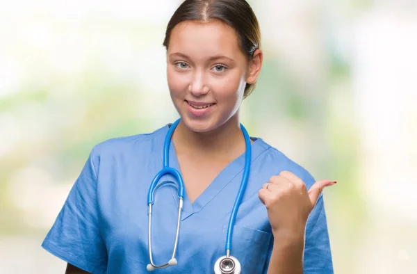 年轻的高加索医生妇女穿着医疗制服在孤立的背景微笑与愉快的面孔看和指向边用拇指 — 图库照片