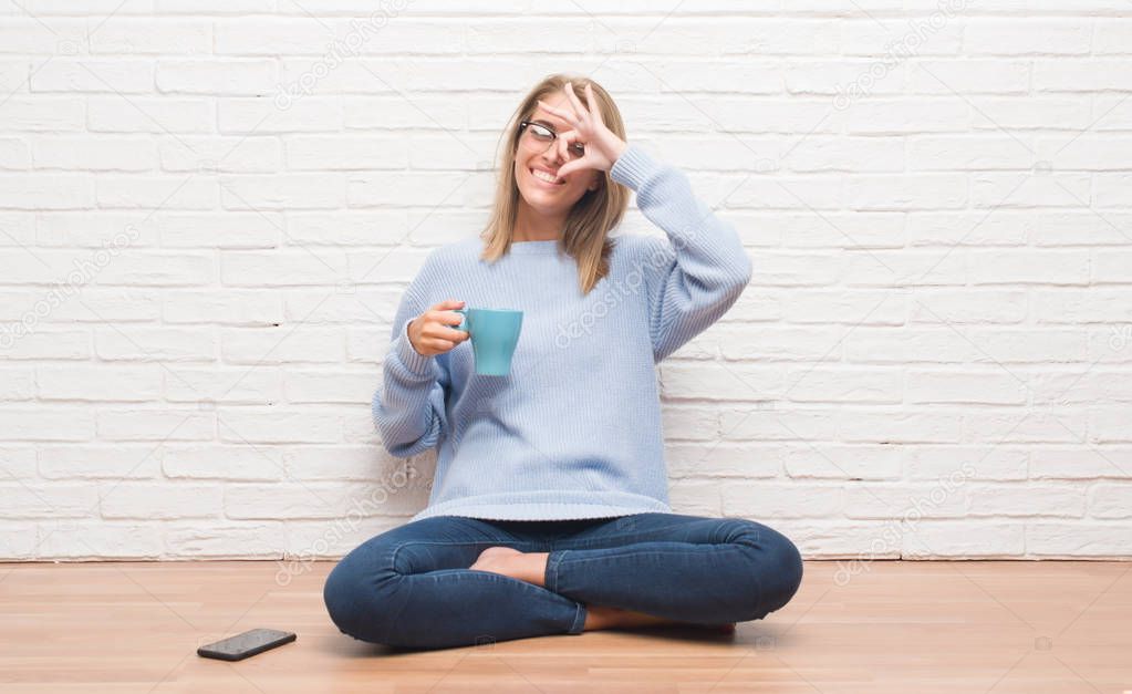 Beautiful young woman sitting on the floor driking cup of coffee at home with happy face smiling doing ok sign with hand on eye looking through fingers