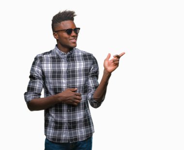 Kameraya bakarak yan el ve parmak ile işaret eden genç Afro-Amerikan adam yüzünde kocaman bir gülümseme ile izole arka plan üzerinde güneş gözlüğü takıyor.