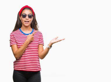 Genç güzel Arap kadın hayrete ve el ile sunulması ve parmak ile işaret ederken kameraya gülümseyen izole arka plan üzerinde güneş gözlüğü takıyor.