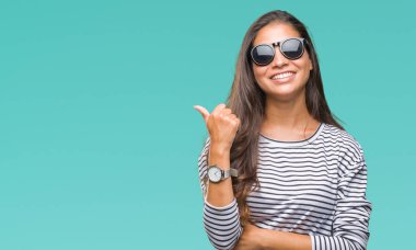 Genç güzel Arap kadın arıyorum ve başparmak yanında yukarıyı mutlu yüz ile gülümseyen izole arka plan üzerinde güneş gözlüğü takıyor.