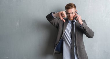 Genç redhead iş adamı kızgın yüzü, eksi işareti aşağı antipati başparmak ile gösterilen, ret kavramı ile telefonda konuşurken gri grunge duvarın üzerinden