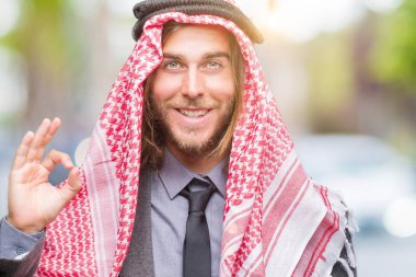 Genç yakışıklı Arap adam uzun saçlı keffiyeh izole arka plan el ve parmaklar ok işaretiyle yapıyor olumlu gülümseyen üzerinde giyiyor. Başarılı ifade.