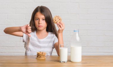 Süt içme ve kızgın yüzü, eksi işareti aşağı antipati başparmak ile gösterilen, ret kavramı ile Çikolatalı bisküvi yemek masada oturan genç İspanyol çocuk