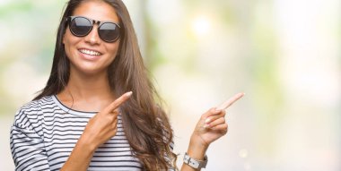 Genç güzel Arap kadın gülümseyerek ve iki elleri ve parmakları ile tarafına işaret kamera bakarak izole arka plan üzerinde güneş gözlüğü takıyor.