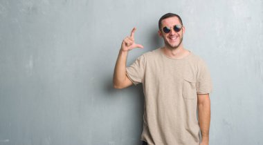 Beyaz genç gülümseyen güneş gözlüğü ve boyutu işaret parmaklı ararken ve kamera işi el ile kendinden emin eller gri grunge duvarın üzerinden. Ölçü kavramı.