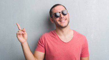 El ve parmak ile tarafına işaret çok mutlu güneş gözlüğü takıyor gri grunge duvarın üzerinden beyaz delikanlı