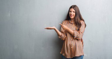 Genç esmer kadın hayrete ve el ile sunulması ve parmak ile işaret ederken kameraya gülümseyen grunge gri duvarın üzerinden.