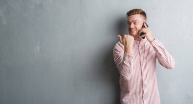 Genç Kızıl saçlı adam işaret ve başparmak kadar mutlu yüz gülümseyen tarafı ile gösterilen telefonda konuşurken gri grunge duvarın üzerinden