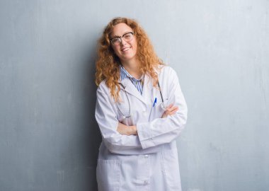Genç redhead doktor kadın kameraya bakarak çapraz silah ile gülümseyerek mutlu bir kat yüz giyen gri grunge duvarın üzerinden. Olumlu bir kişi.