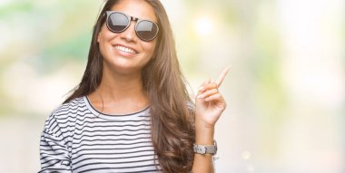 Kameraya bakarak yan el ve parmak ile işaret yüzünde kocaman bir gülümseme ile izole arka plan üzerinde güneş gözlüğü takmış genç güzel Arap kadın.