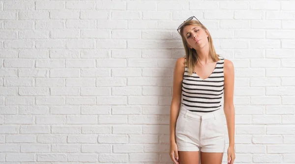 Young beautiful blonde woman over white brick wall making fish face with lips, crazy and comical gesture. Funny expression.