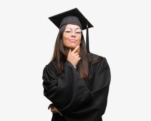 年轻的西班牙裔妇女穿着毕业帽和制服看着自信的相机与交叉的手臂和手举起下巴 积极思考 — 图库照片