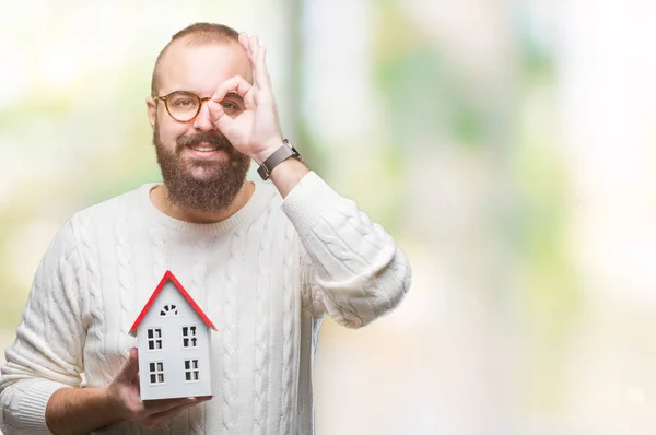Young caucasian real state agent man holding house isolated background with happy face smiling doing ok sign with hand on eye looking through fingers